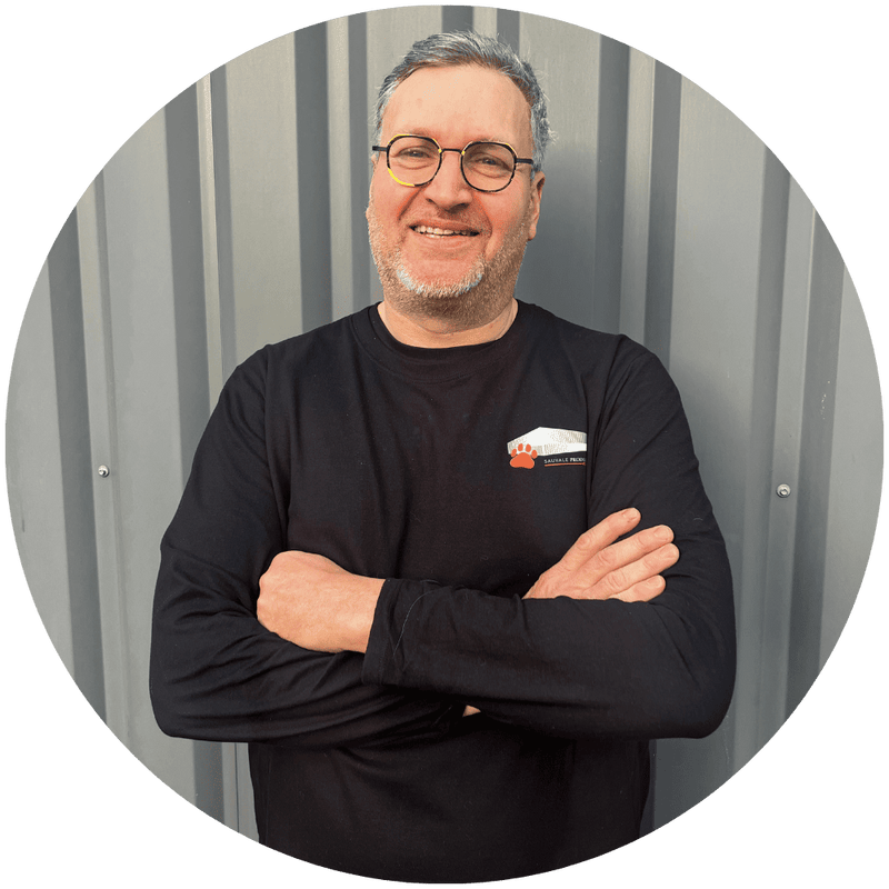 Christophe, préparateur de commandes chez Stan.bio, les bras croisés et souriant, portant un t-shirt noir avec le logo Sauvale Premium, devant une façade en métal gris