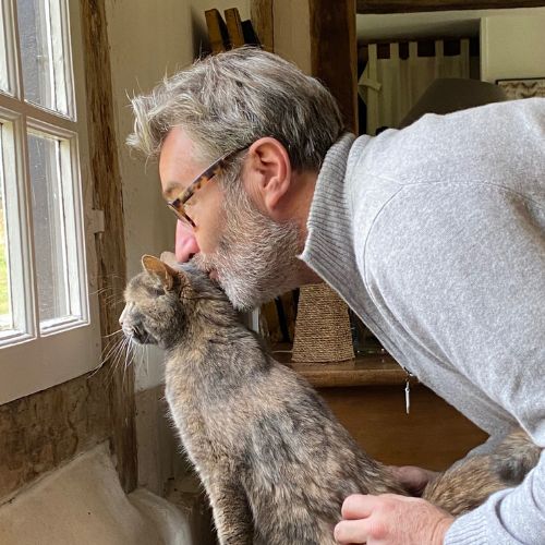 Stan de Vion fondateur de Stan Bio câlinant tendrement son chat tabby gris près d'une fenêtre dans sa maison de campagne